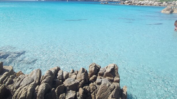 Un paysage côtier caractérisé par une eau turquoise et limpide, des formations rocheuses et une colline qui se dessine à l’horizon.