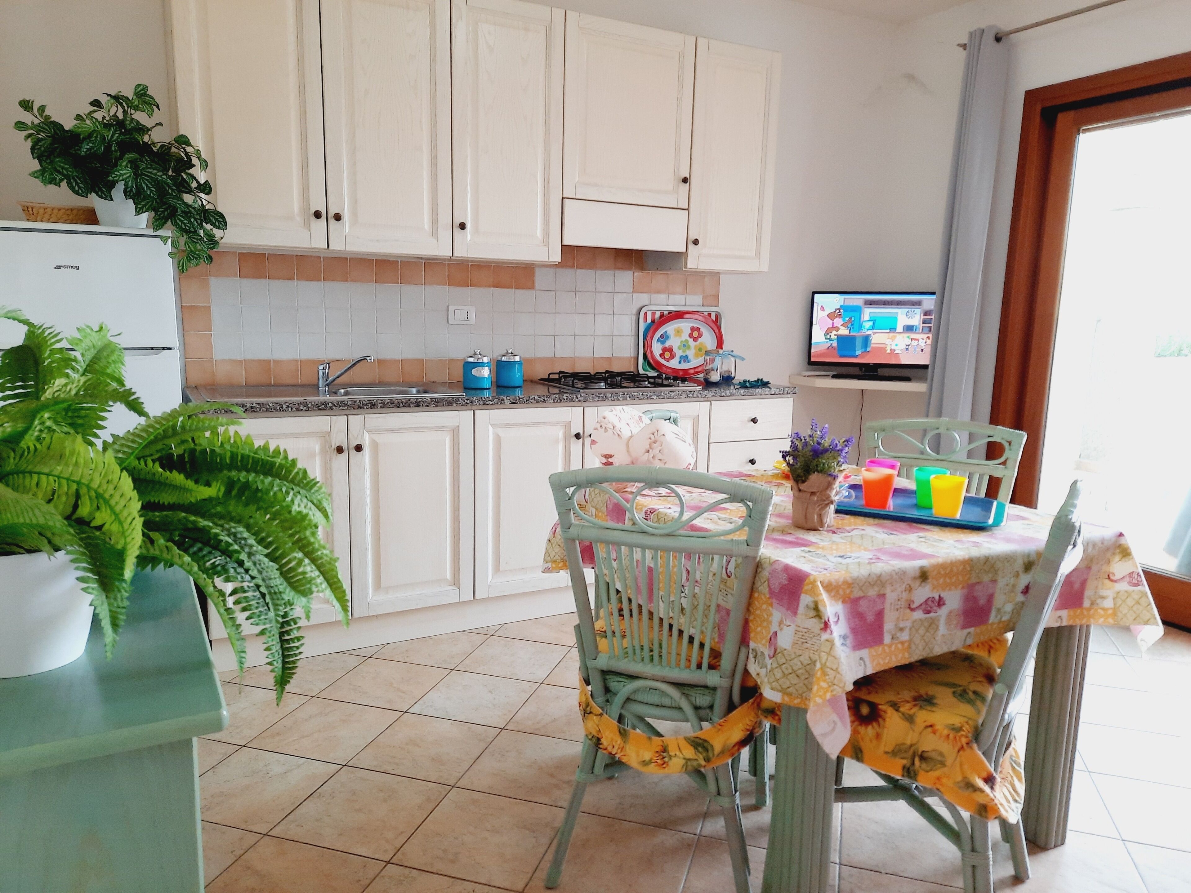 A kitchen with white cabinets, a tiled backsplash, a sink, and a stove. There is a dining table with chairs and a TV in the background.