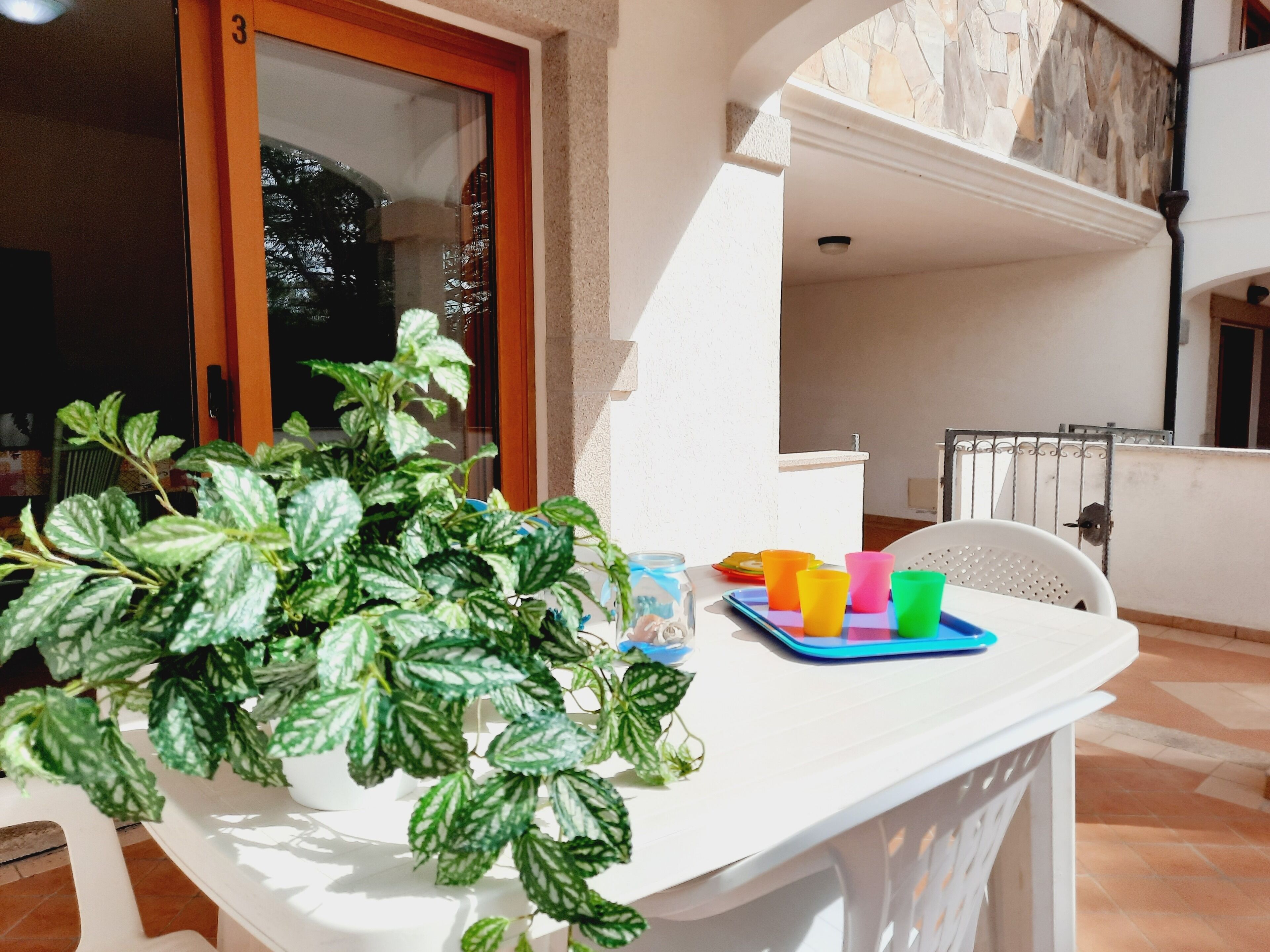 Un balcón con una mesa blanca, una planta en maceta, tazas de colores y una barandilla de metal.