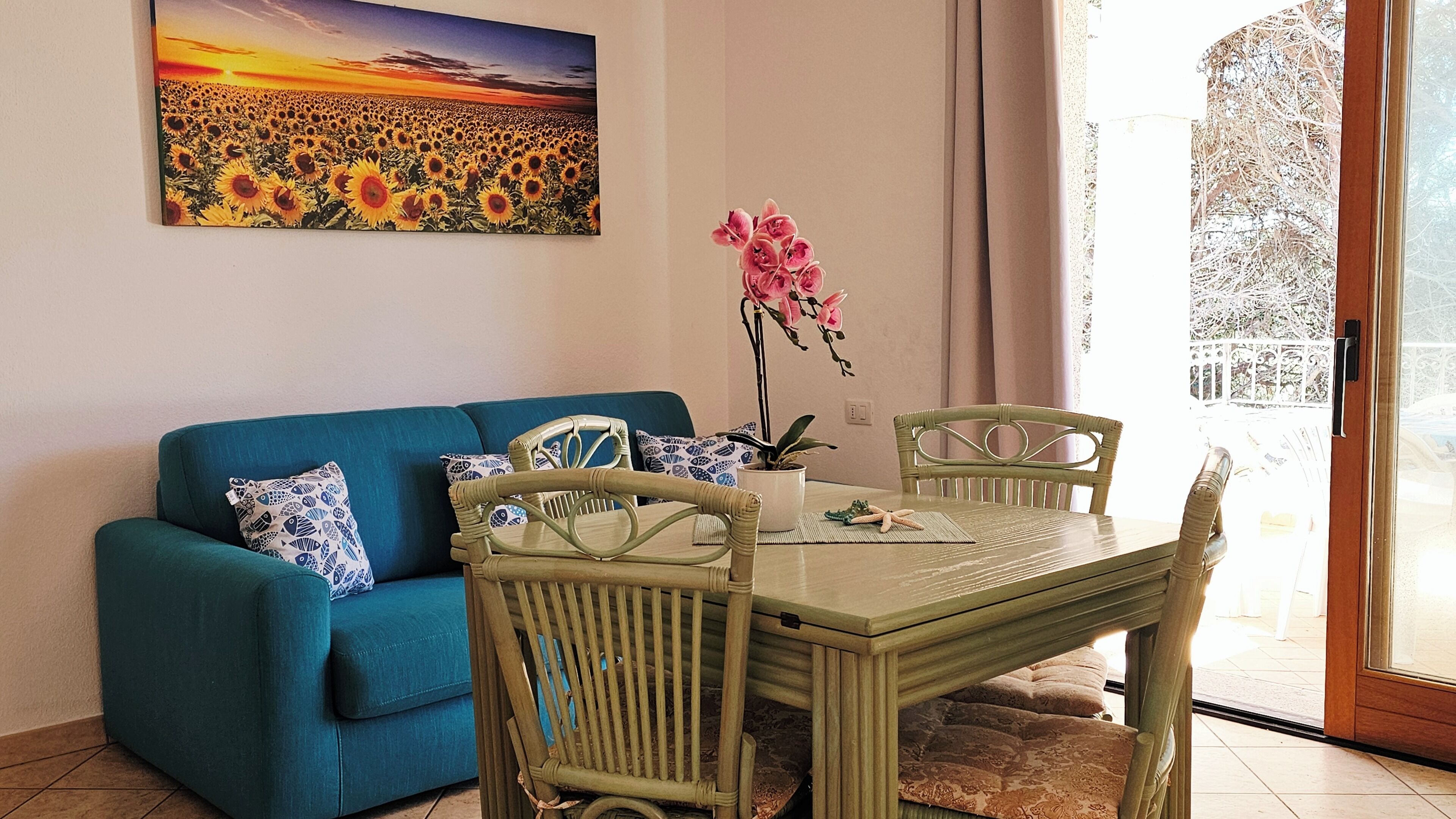 Una sala de estar con un sofá azul, una mesa de comedor con sillas, una orquídea en maceta y un cuadro de girasoles.
