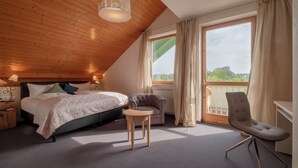 Een slaapkamer met een bed, een stoel, een tafel, een raam met gordijnen en uitzicht op groen.