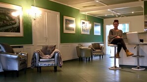 Een kapsalon met groene muren, meerdere zithoeken, en een persoon zittend op een hoge stoel.