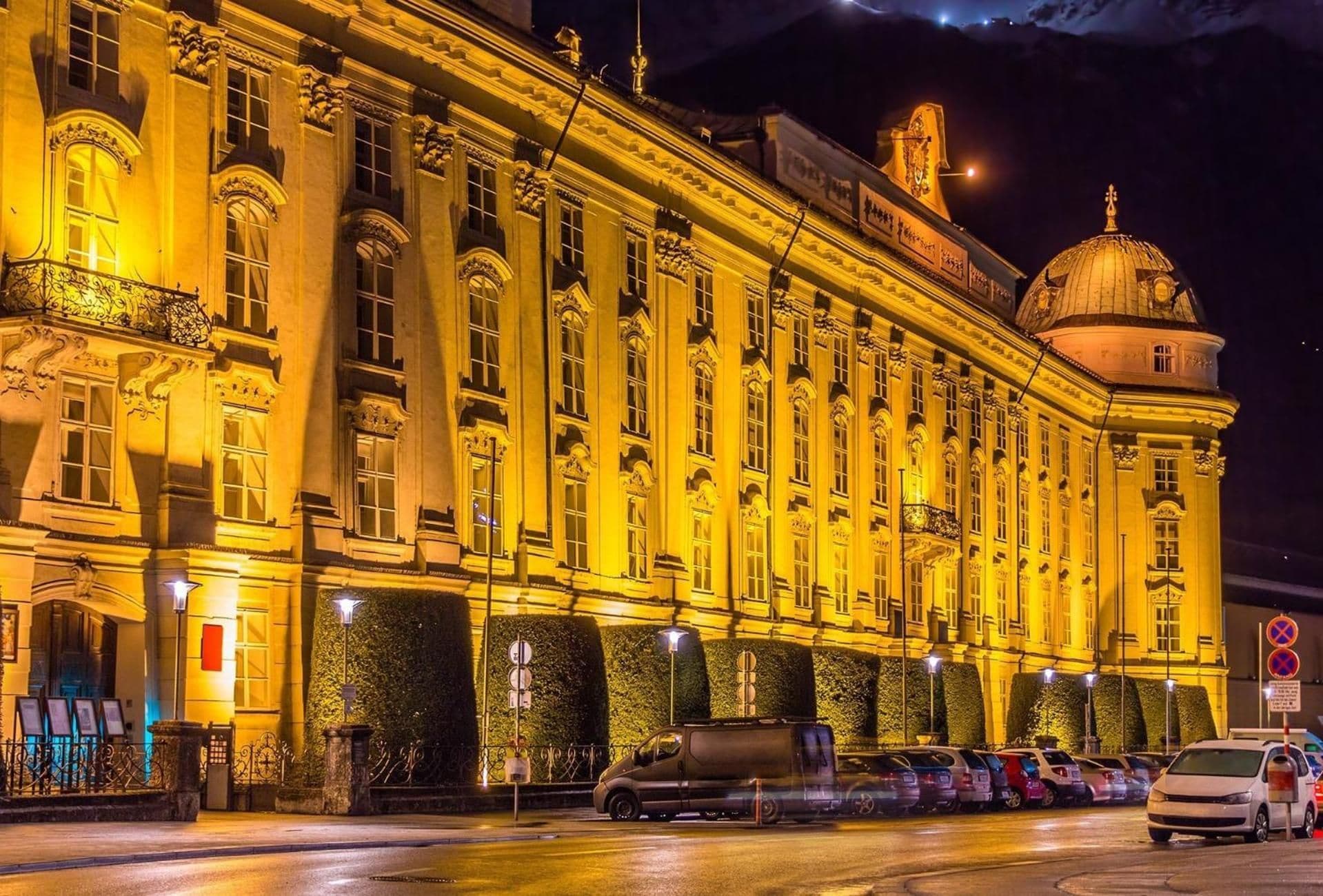 A large, historic building with a prominent dome and classical architecture, featuring a well-lit facade and a parking area in front.