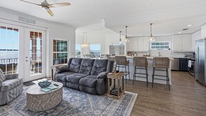 Een ruime woonkamer met een grijze bank, een ronde salontafel en een vloerkleed met dessin. Er is een balkon met uitzicht op het water, en de keuken is zichtbaar met witte kasten en een roestvrijstalen koelkast.