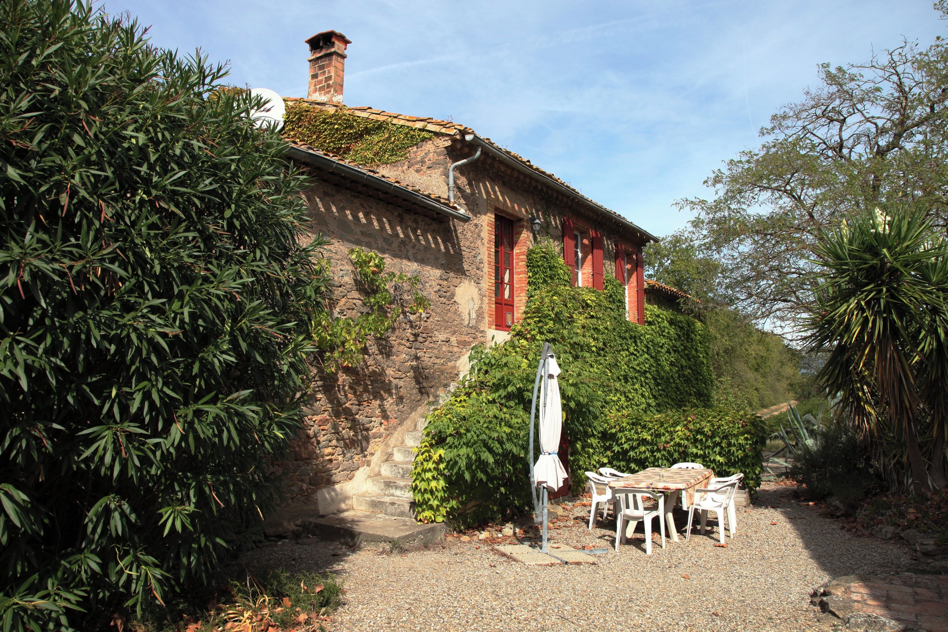 Un bâtiment rustique en pierre, doté de volets rouges, d’une terrasse en gravier et d’une table avec des chaises.