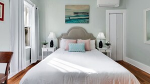 A bedroom with a bed, two bedside tables with lamps, a painting of a pier, an air conditioning unit, and a door.