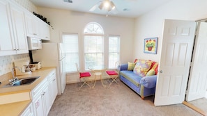 A compact kitchen with white cabinets, a refrigerator, and a coffee maker. There is a dining area with a table and chairs, and a blue sofa with colorful cushions.