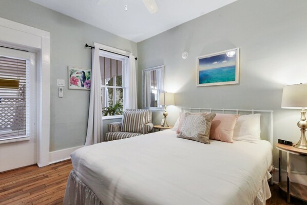 A bedroom with a large bed, a window with curtains, a bedside table with a lamp, and a painting on the wall.