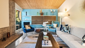 Un salon moderne avec un plafond en bois, des murs d’accent bleu, un canapé gris, des fauteuils bleus et une table basse avec des livres.