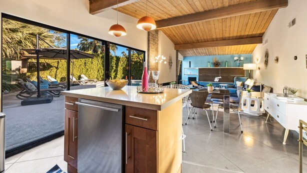 Une cuisine moderne dotée d’un îlot central, de poutres en bois au plafond et de grandes fenêtres donnant sur un patio aménagé avec des sièges d’extérieur.