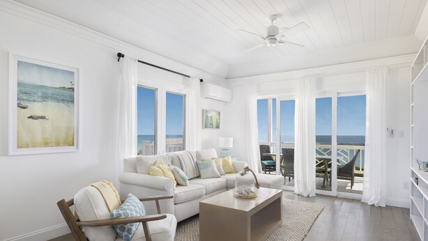 A spacious living room with a white sofa, a wooden chair, a coffee table, and a view of the ocean through large windows. - Condo w/Views, Beach, Htd Pool, Golf Cart, Kayaks, Gym, Pickleball Court! (Governors Harbour)