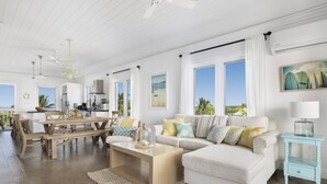 A spacious living room with a white sofa, a wooden dining table, and a kitchen in the background. - Condo w/Views, Beach, Htd Pool, Golf Cart, Kayaks, Gym, Pickleball Court! (Governors Harbour)