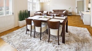 Ein modernes Wohnzimmer mit Esstisch, Stühlen, einer Couch und einem Couchtisch. Ein großes Fenster, eine Pflanze und ein Kronleuchter sind ebenfalls vorhanden.