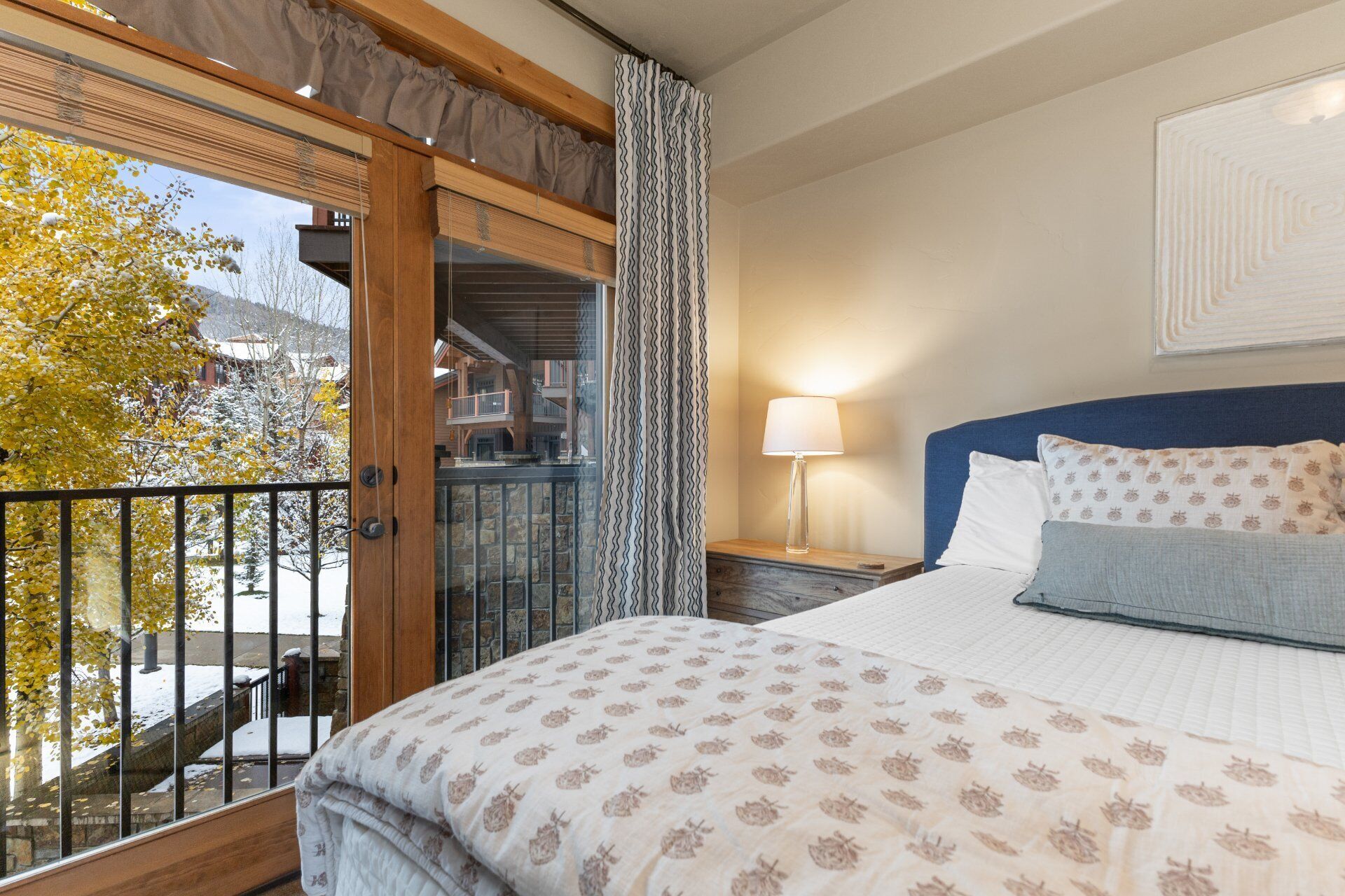 A bedroom with a bed, a nightstand, a lamp, a window with curtains, and a view of a building and trees outside.