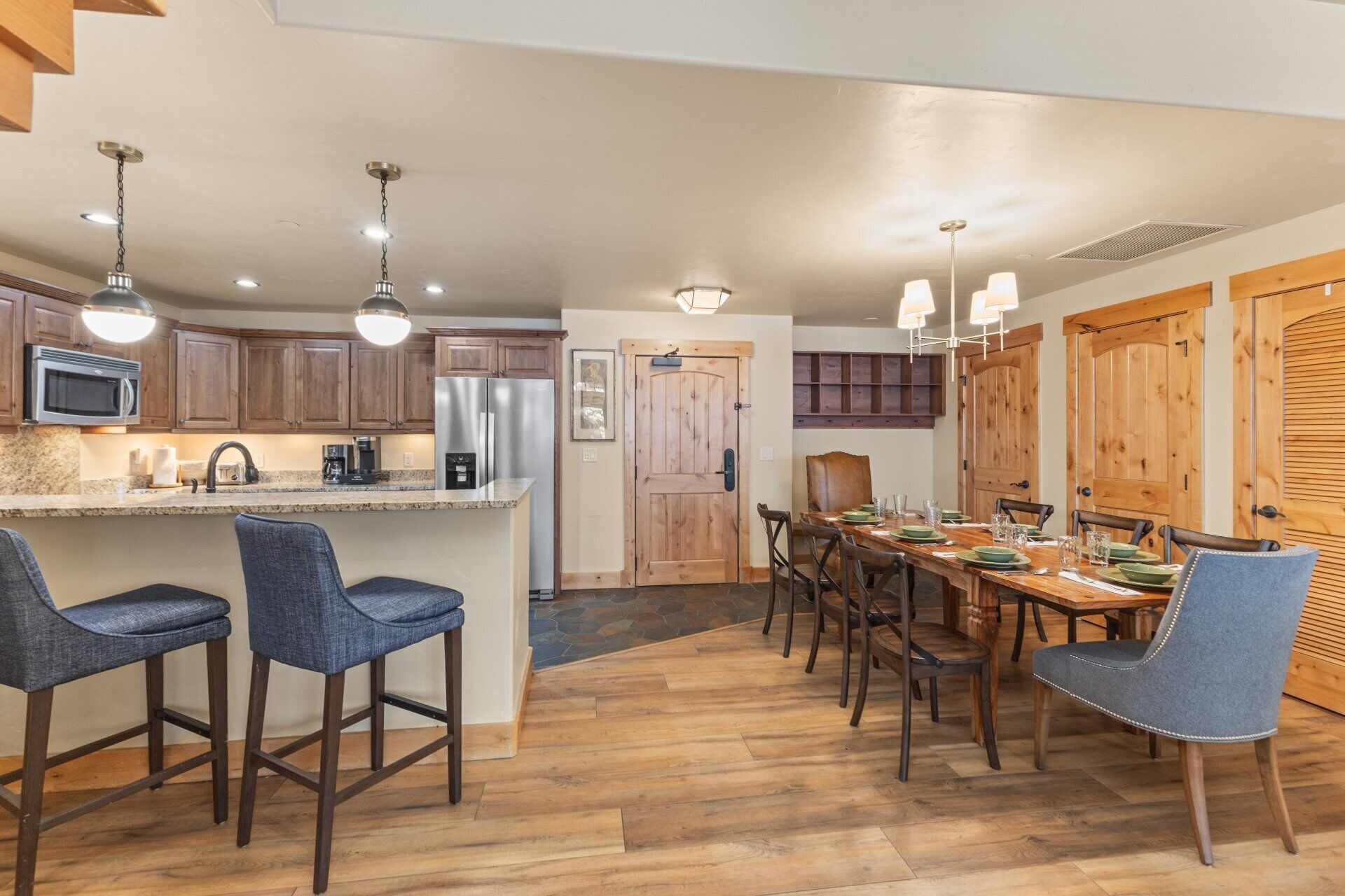 A modern kitchen with a central island, bar stools, a dining table with chairs, wooden cabinets, and a refrigerator.