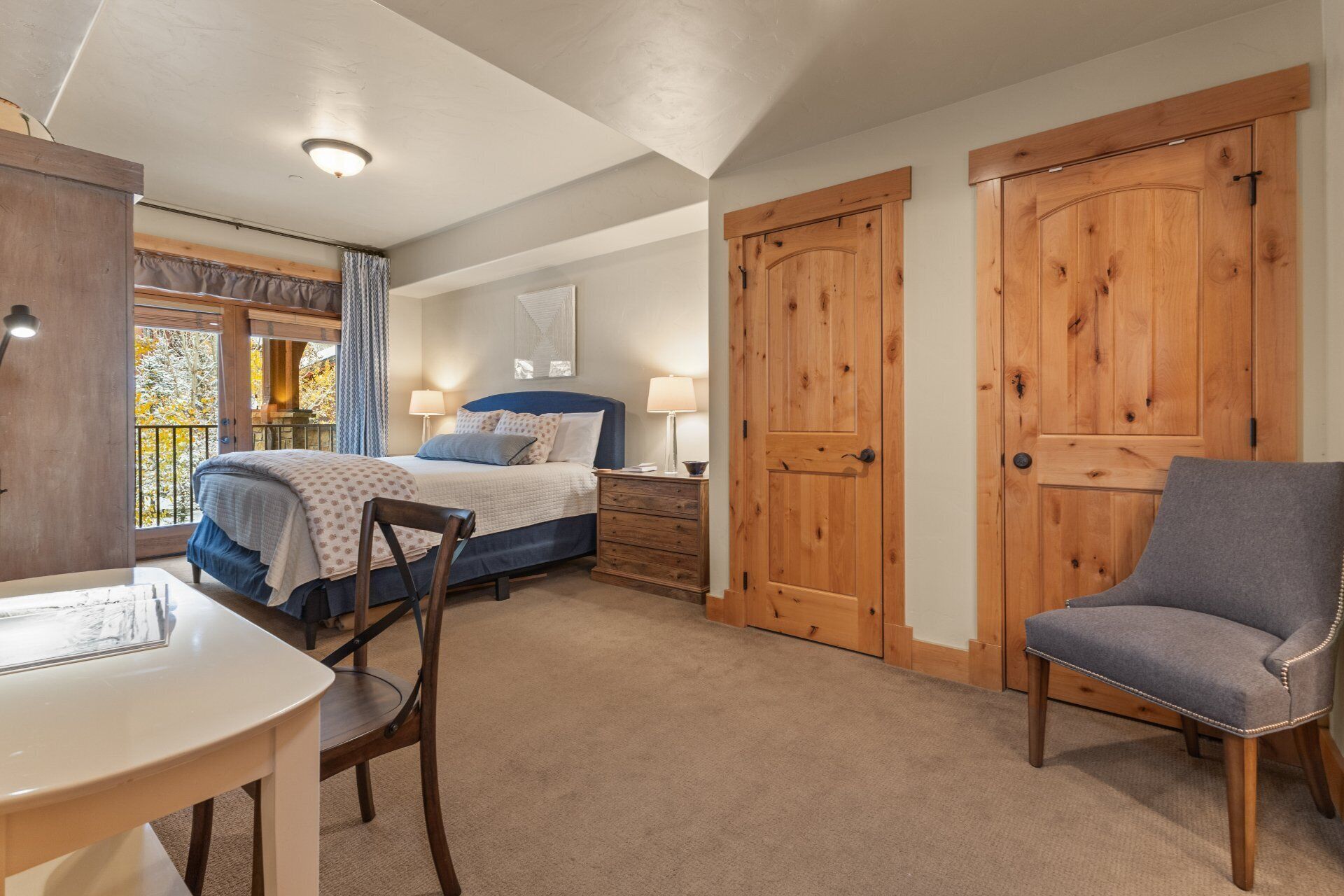 A bedroom with a bed, bedside tables, a chair, and a desk. There are two wooden doors and a window with curtains.