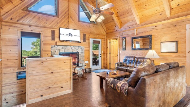 A spacious wooden cabin interior with a stone fireplace, a ceiling fan, and a large window offering a view of the outdoors.