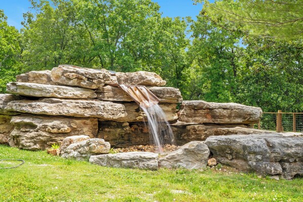 A natural rock formation with a waterfall in a grassy area, surrounded by trees.