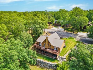 A two-story log cabin with a deck and a stone wall, surrounded by dense forest.