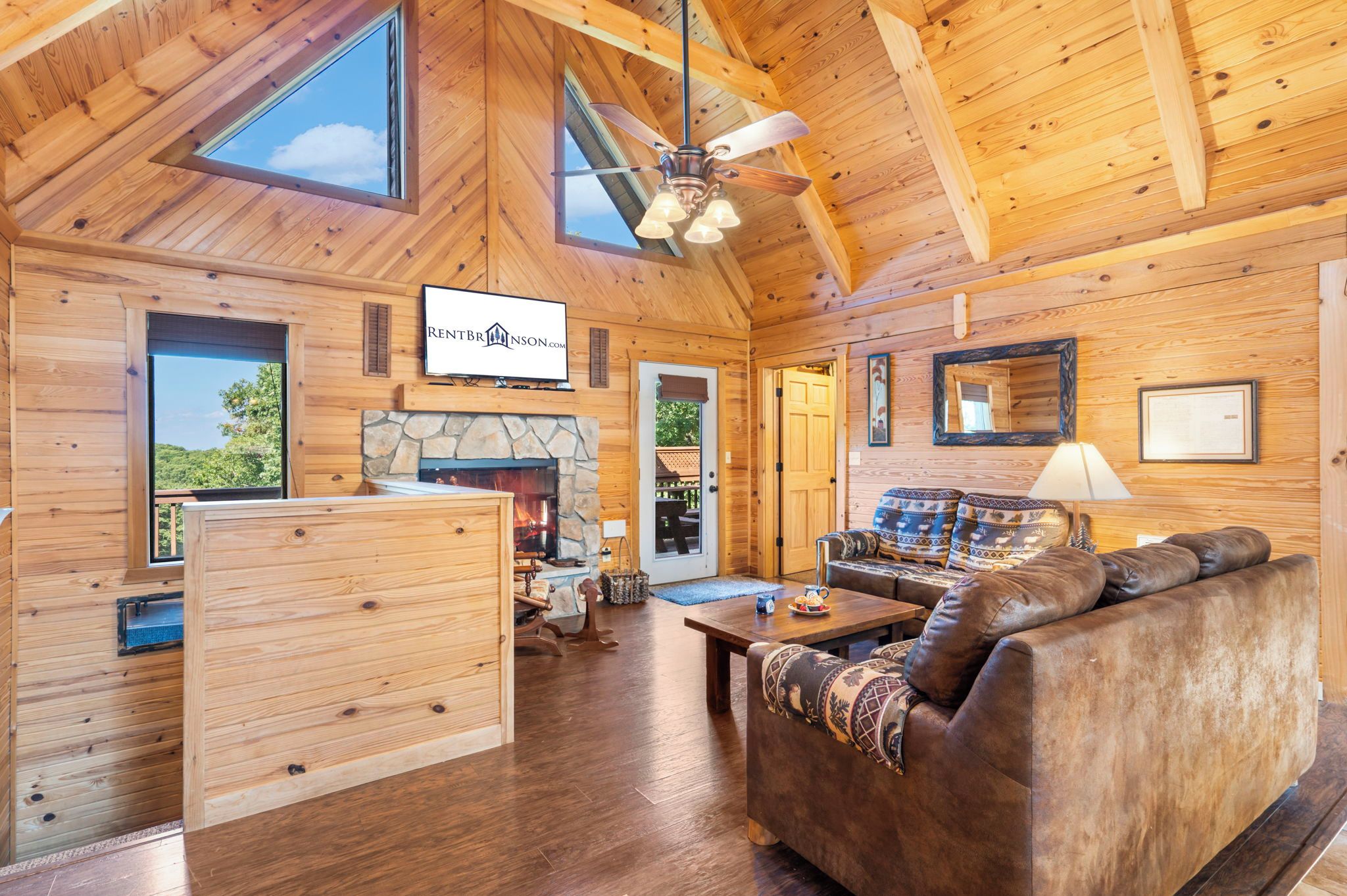 A cozy cabin interior with a stone fireplace, wooden walls, a ceiling fan, and a television mounted above the fireplace.