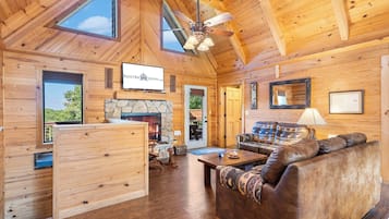 L’intérieur chaleureux d’un chalet avec une cheminée en pierre, des murs en bois, un ventilateur de plafond et un téléviseur fixé au-dessus de la cheminée.