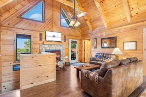 A cozy cabin interior with a stone fireplace, wooden walls, a ceiling fan, and a television mounted above the fireplace. - Elegant 2700 Sq Ft True Log Cabin Near Big Cedar_Private Hot Tub_Game Room_Pool Table_Best View (Ridgedale)