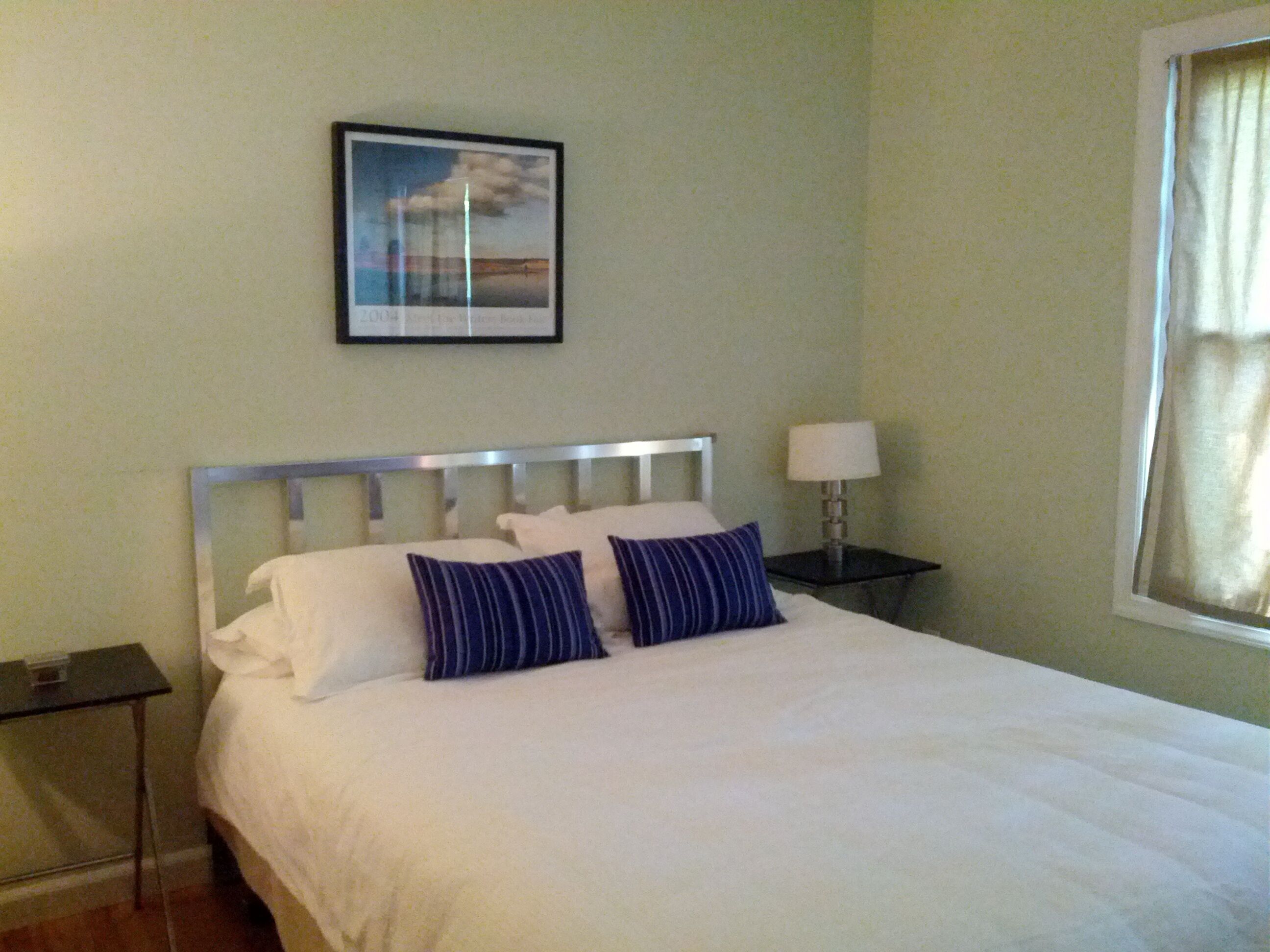 A neatly made bed with white linens and two blue striped pillows, a bedside table with a lamp, and a framed picture on the wall.