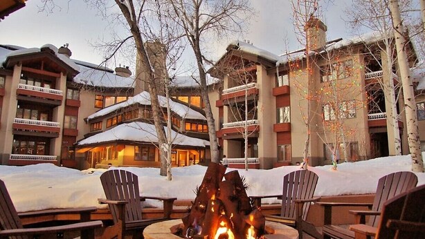 Un chalet doté d’un foyer extérieur et de sièges en plein air, entouré de jardins et de bâtiments recouverts de neige et dotés de balcons.