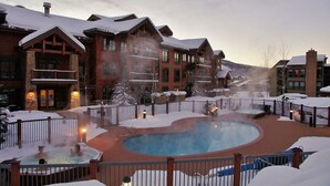Un espace extérieur avec une piscine recouverte de neige et un jacuzzi, entouré de bâtiments en bois dotés de balcons.