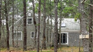 A rustic cabin surrounded by tall trees, with a visible door and a number '75' sign on a tree. - Newly Updated Spacious Home in Quiet Wooded Area (1246) (Wellfleet)