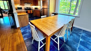 A modern kitchen with wooden cabinets, a stainless steel refrigerator, and a dining area with a wooden table and white chairs. - Newly Updated Spacious Home in Quiet Wooded Area (1246) (Wellfleet)