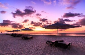 A beach with umbrellas and lounge chairs, a sunset in the background, and a calm sea.