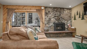 A cozy living room with a stone fireplace, a large window with curtains, and a view of a snowy building outside. - Easy Walk to Base Area Activities - Cozy Condo + Large Outdoor Hot Tub (Steamboat Springs)