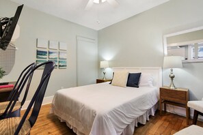 Ein ordentlich eingerichtetes Schlafzimmer mit einem Bett, zwei Nachttischen mit Lampen, einem Sessel und einer Wand mit gerahmten Bildern.