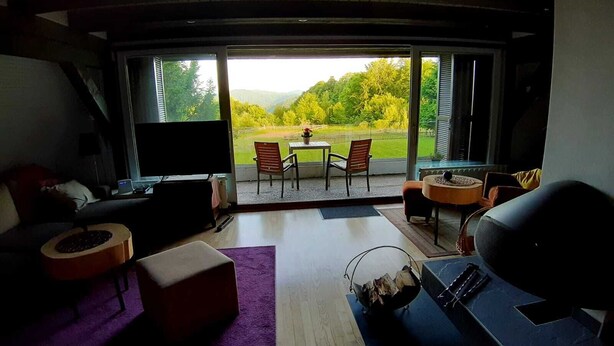 A modern living room with a large TV, comfortable seating, a coffee table, and a view of a green landscape through large windows.