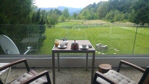 A balcony with a table set for tea, overlooking a grassy field and forested hills.