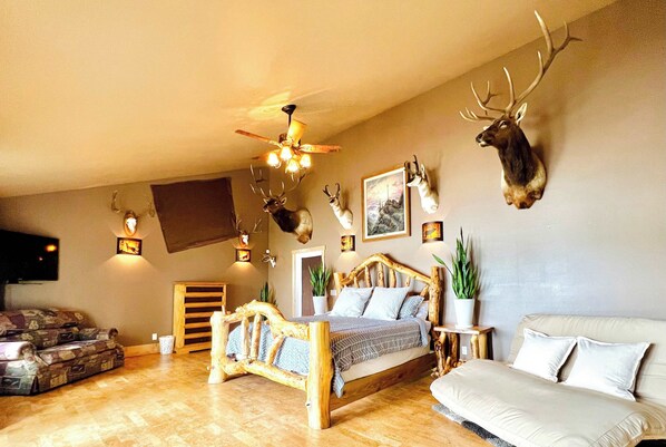 A bedroom with a wooden bed, a ceiling fan, mounted deer heads, and a TV on the wall.