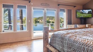 A bedroom with a large bed, a television, and a view of a lake through glass doors. - Amazing Flathead River Location Gunner River Lodge (Bigfork)