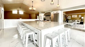 A modern kitchen with a large island, white bar stools, pendant lighting, and a clean, white interior. - Amazing Flathead River Location Gunner River Lodge (Bigfork)