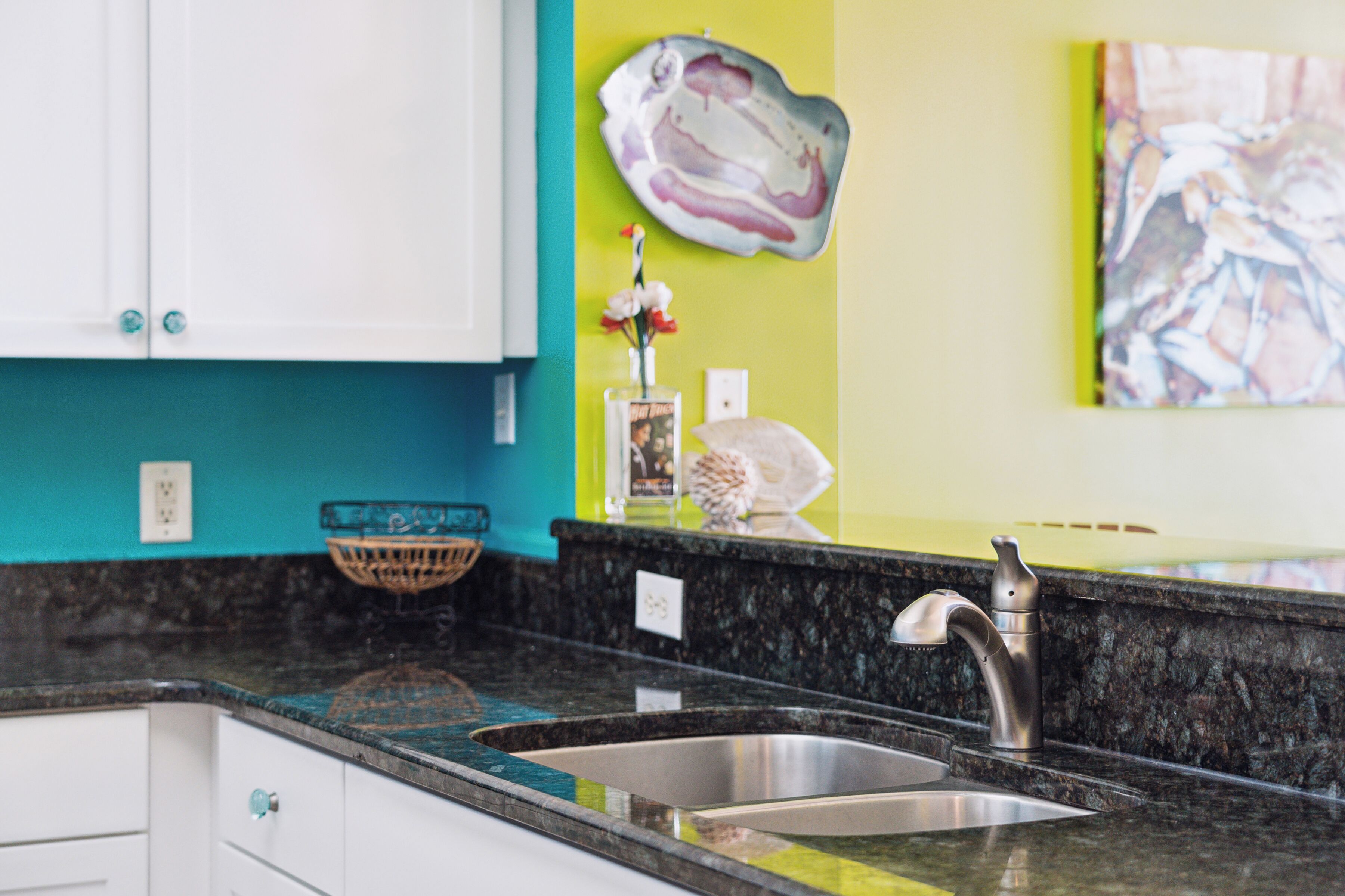Eine Küche mit einem Edelstahl-Spülbecken, einer schwarzen Granit Arbeitsplatte, einem weißen Schrank und einer türkisblauen Wand.