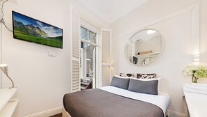 Ein Schlafzimmer mit einem Bett, einem Nachttisch, einem Spiegel, einem Fernseher und einem Fenster mit Rollläden.