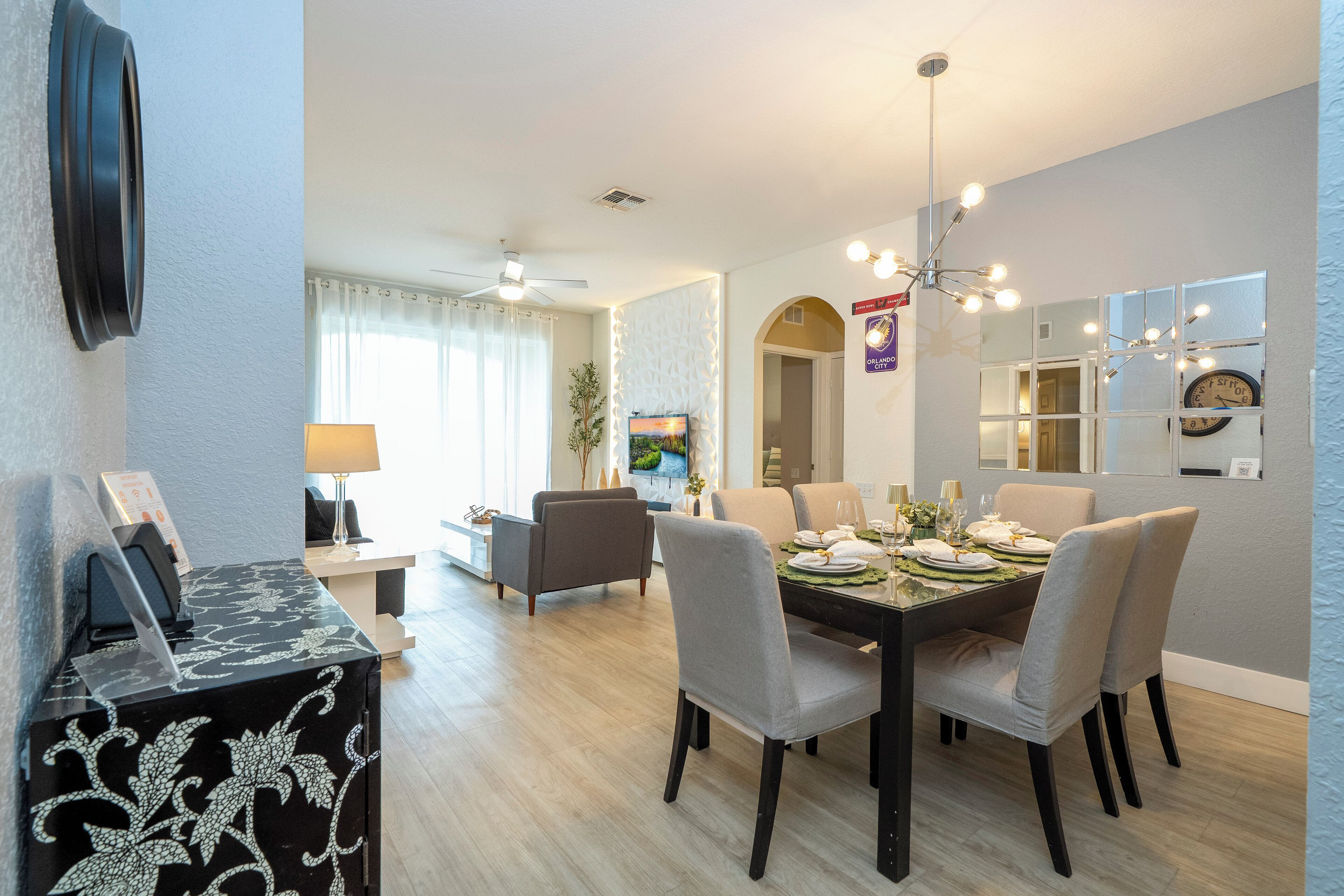 Un coin repas avec une table ronde dressée pour quatre personnes, entourée de six chaises. Un ventilateur de plafond se trouve au-dessus de la table, et une horloge murale est fixée au mur, à droite.