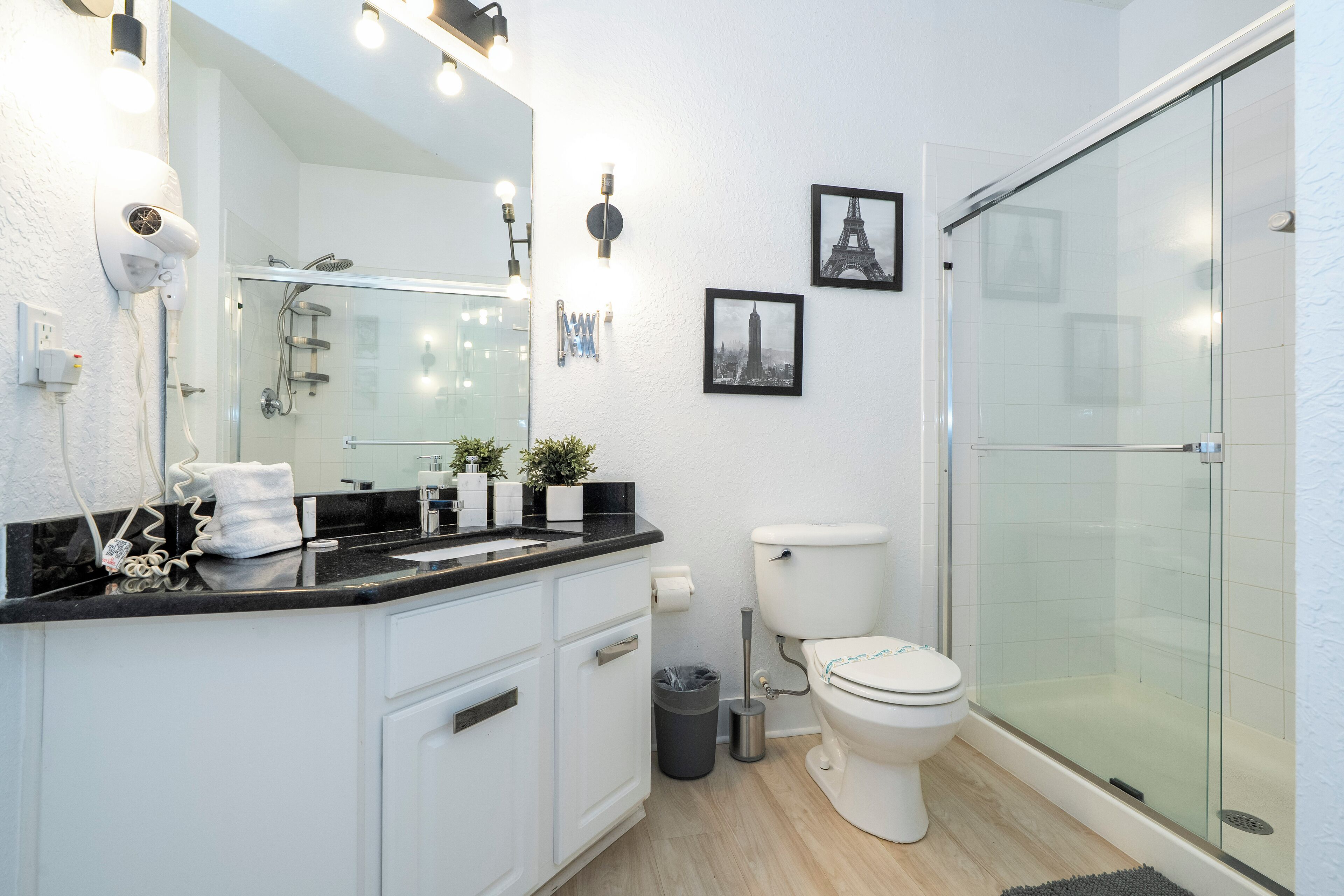 Et moderne badeværelse med en brusekabine i glas, et hvidt toilet og en sort bordplade med et spejl og lys.