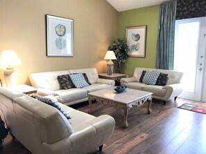 A living room with a beige sofa set, a coffee table, and two side tables with lamps. There are framed botanical prints on the walls.