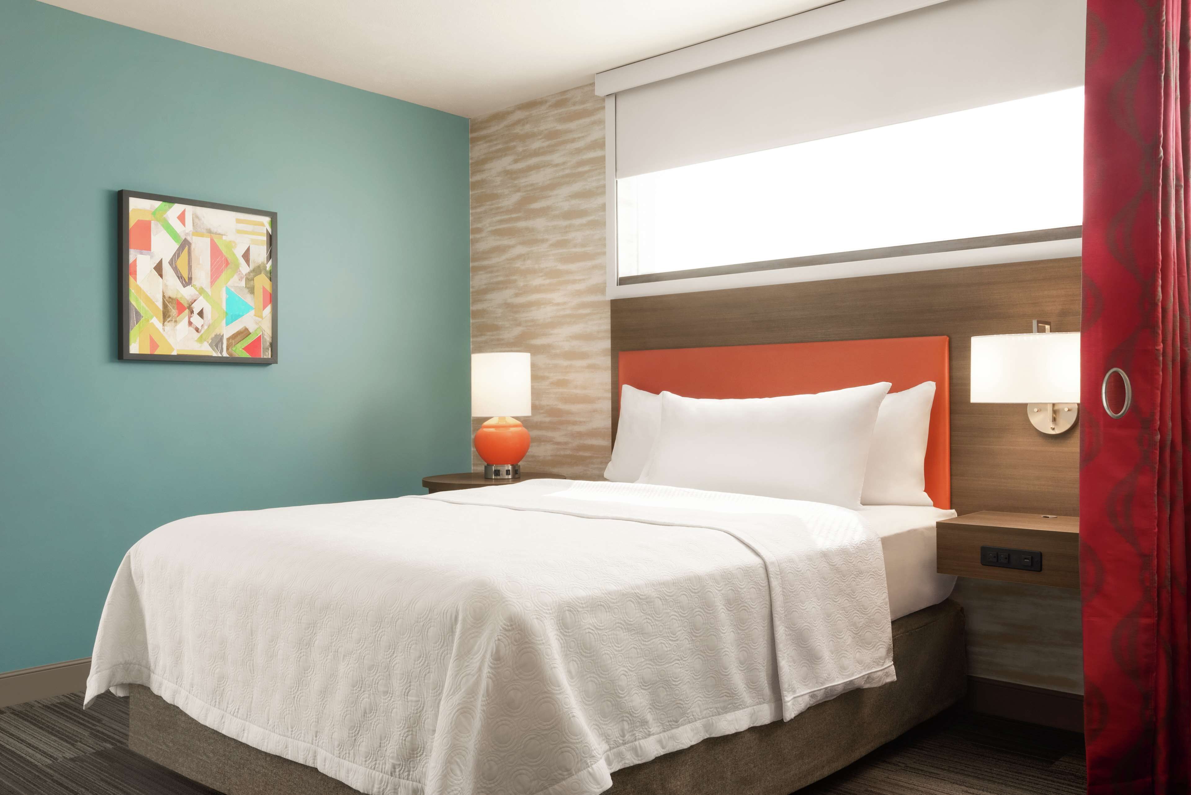 A hotel room with a bed, a nightstand with a lamp, a wall art piece, and a window with curtains.