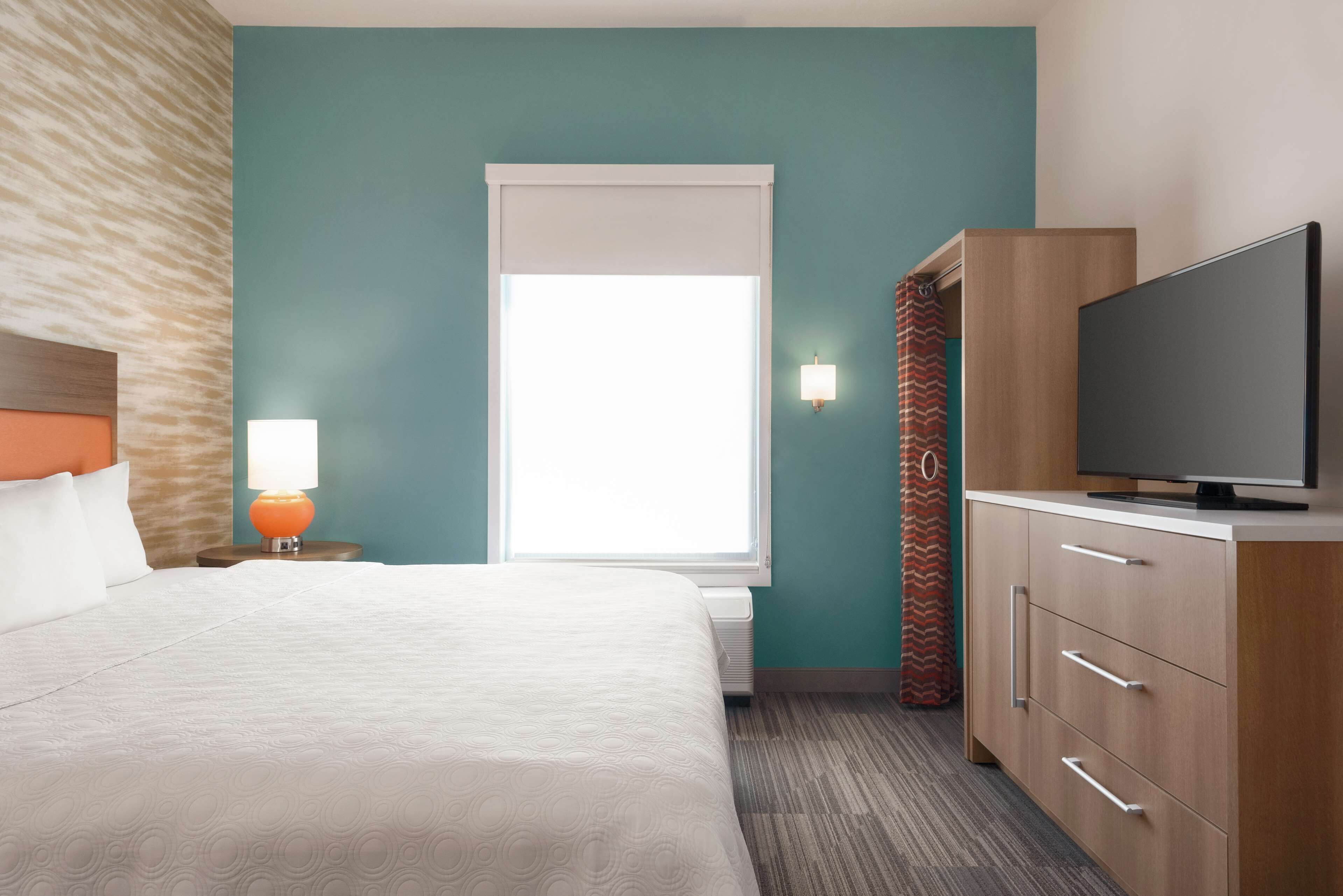 Quarto de hotel com cama, televisão, mesa de cabeceira com abajur, janela com cortinas e guarda-roupa.