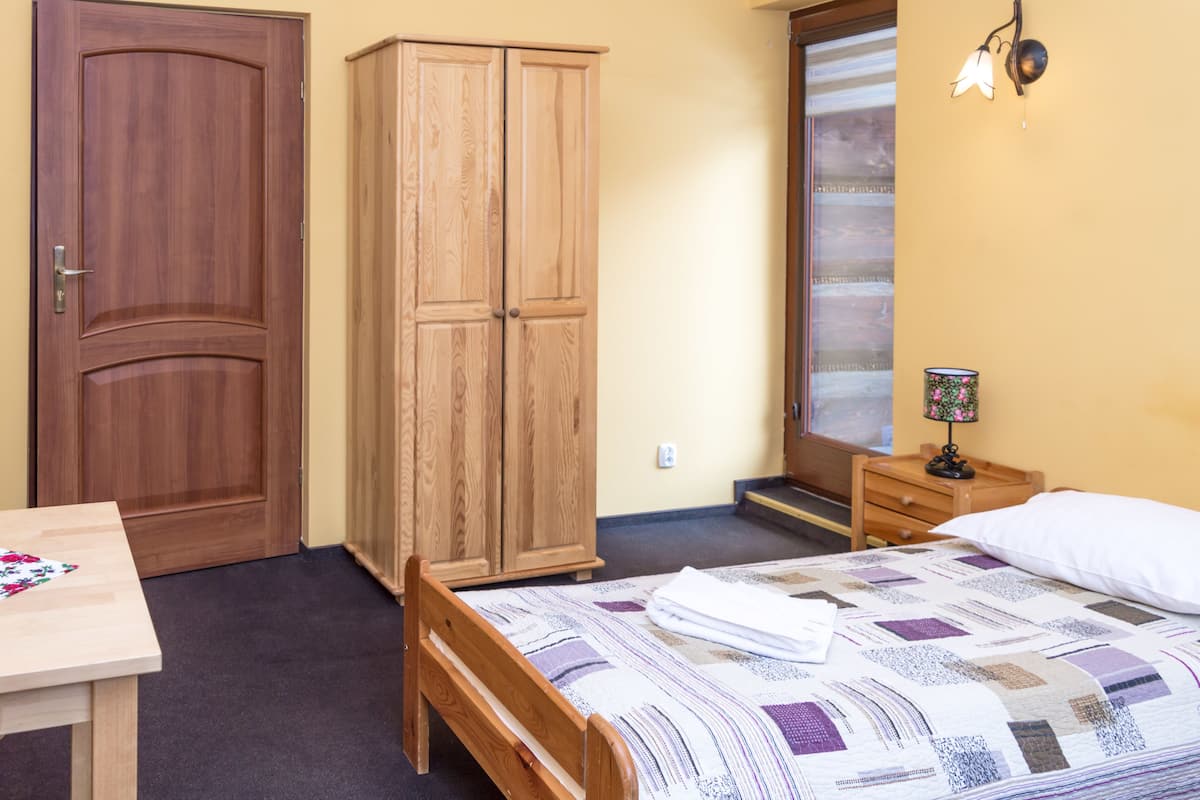 A bedroom with a wooden bed, a wooden wardrobe, a small bedside table with a lamp, and a window with curtains.