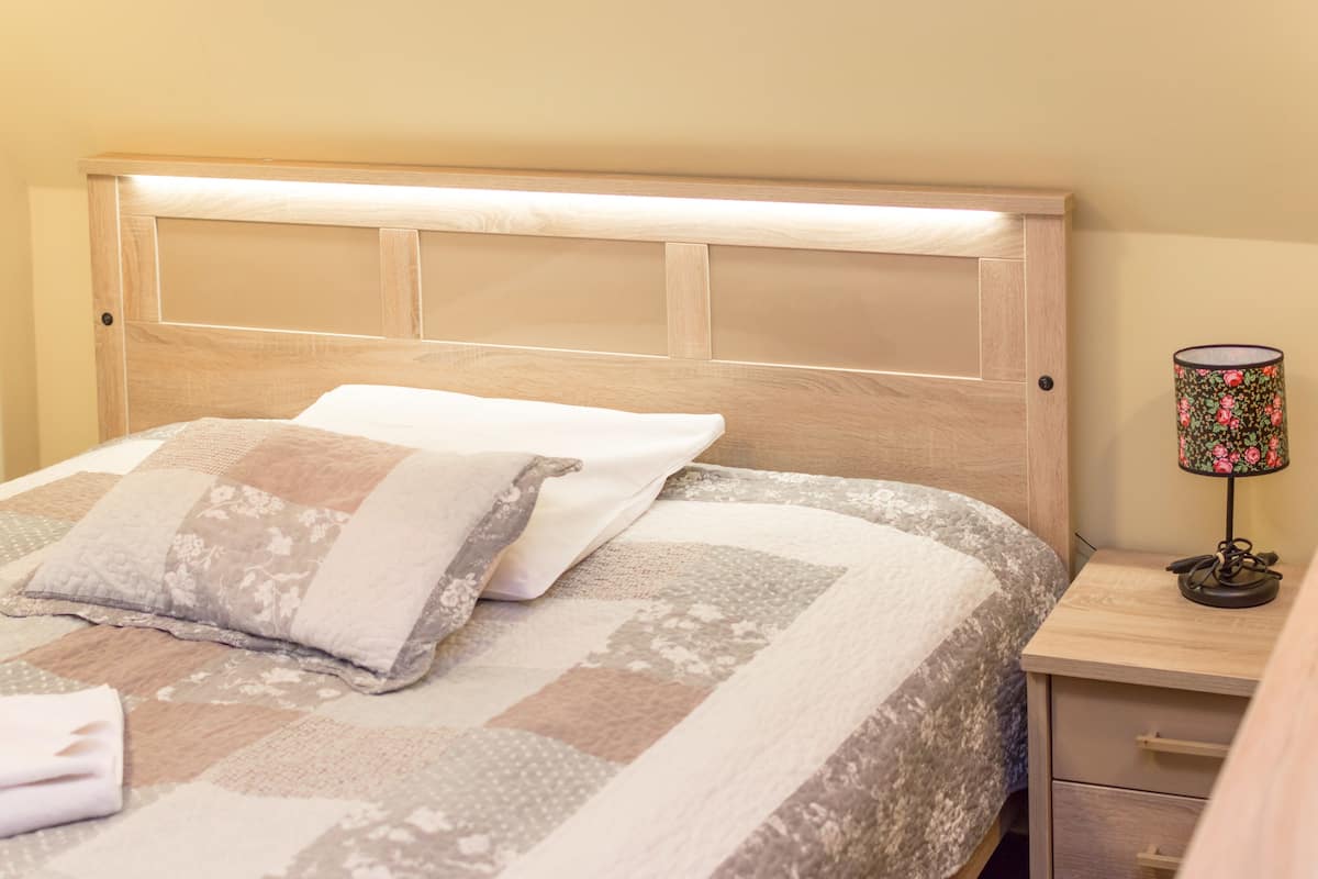 A neatly made bed with a quilted bedspread and two pillows, a wooden headboard, a bedside table with a lamp, and a small drawer.