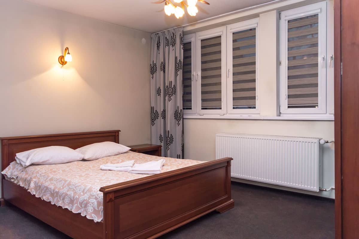 A neatly made bed with a floral bedspread and pillows, a bedside table with a lamp, a window with shutters, and a radiator.