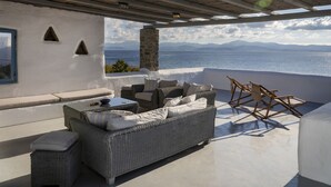 A covered outdoor seating area with a wicker sofa, cushions, and a wooden chair. A stone pillar and a view of the sea and mountains are visible. -  Villa With Swimming Pool In Front Of Almost Private Beach  (PAROS)
